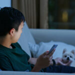 man holds cell phone while petting small dog on the couch