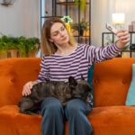woman sits on a couch with her Frenchie dog on a telehealth veterinary visit